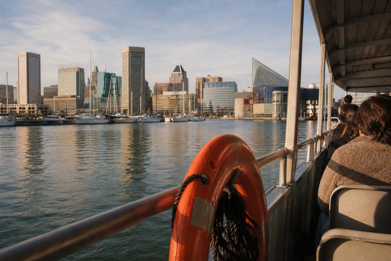 An orange life ring mounted on the rail of a small open harbor shuttle, calm green Inner Harbor water and the softened skyline behind it in early afternoon light