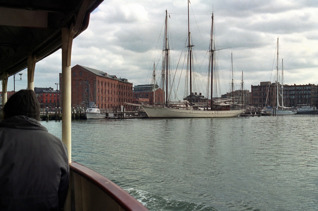 Brick warehouses and tall-ship rigging along the Fells Point waterfront seen from a small passenger boat crossing the Inner Harbor, low afternoon sun on green water