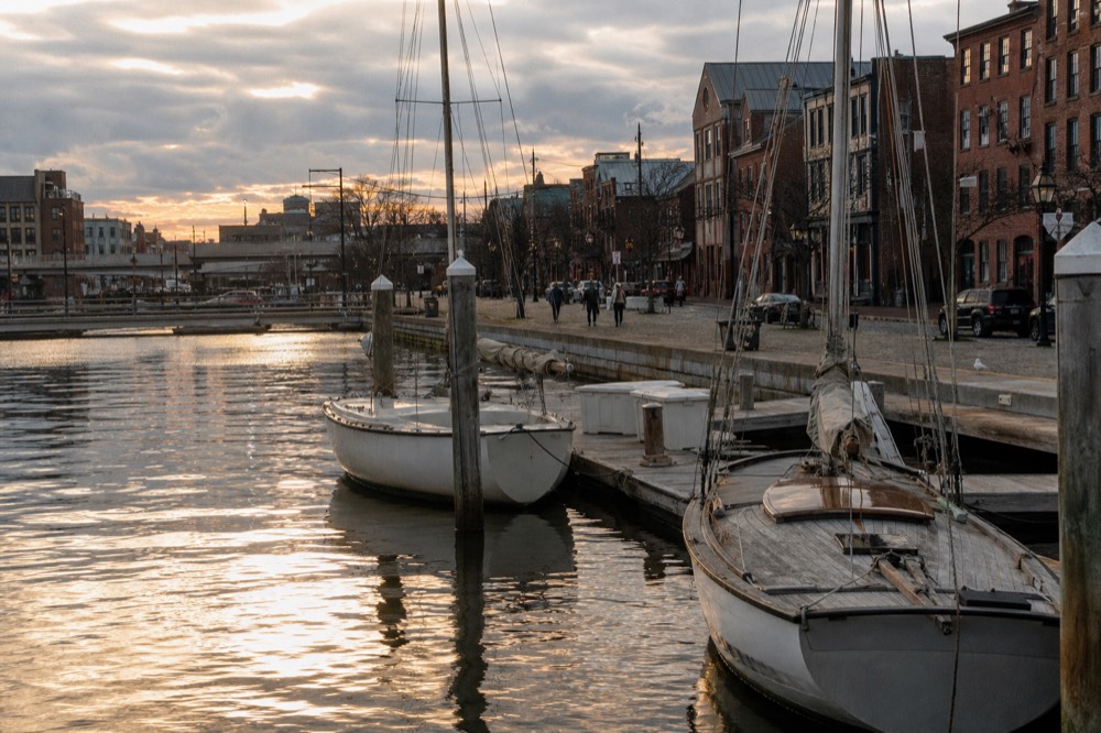 A floating dock with two harbor launches tied up beside numbered piling posts, a cobblestone Fells Point street behind under soft overcast light