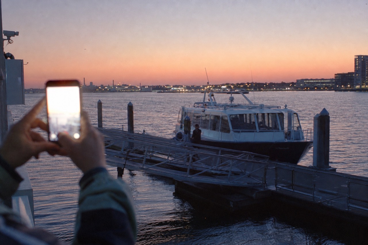 A phone held toward a passenger launch idling at a floating dock, screen glare hiding any detail, brick waterfront blurred behind in bright daylight