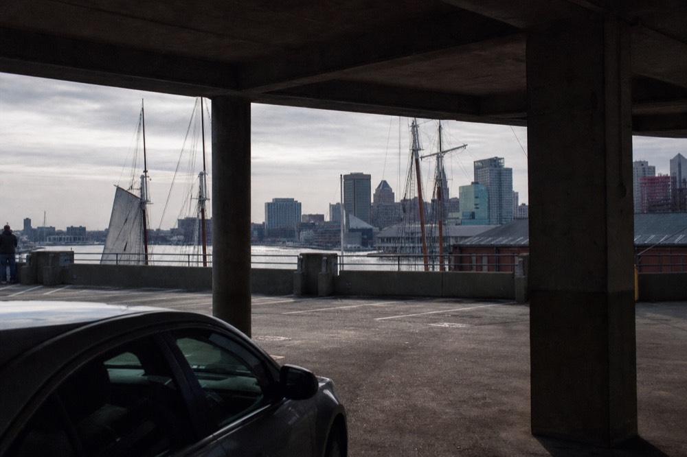 An early-morning upper garage deck nearly empty, the downtown skyline and harbor framed between bare concrete pillars, long shadows across the parked cars