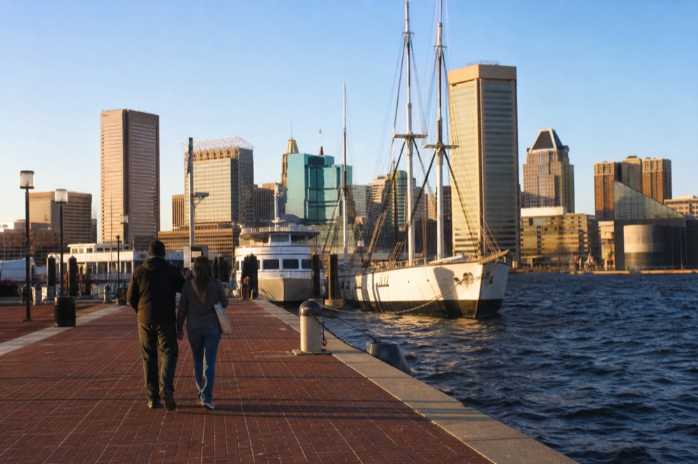 A couple walking a flat brick promenade toward white sightseeing boats moored at the piers, golden evening light on the water and downtown towers behind