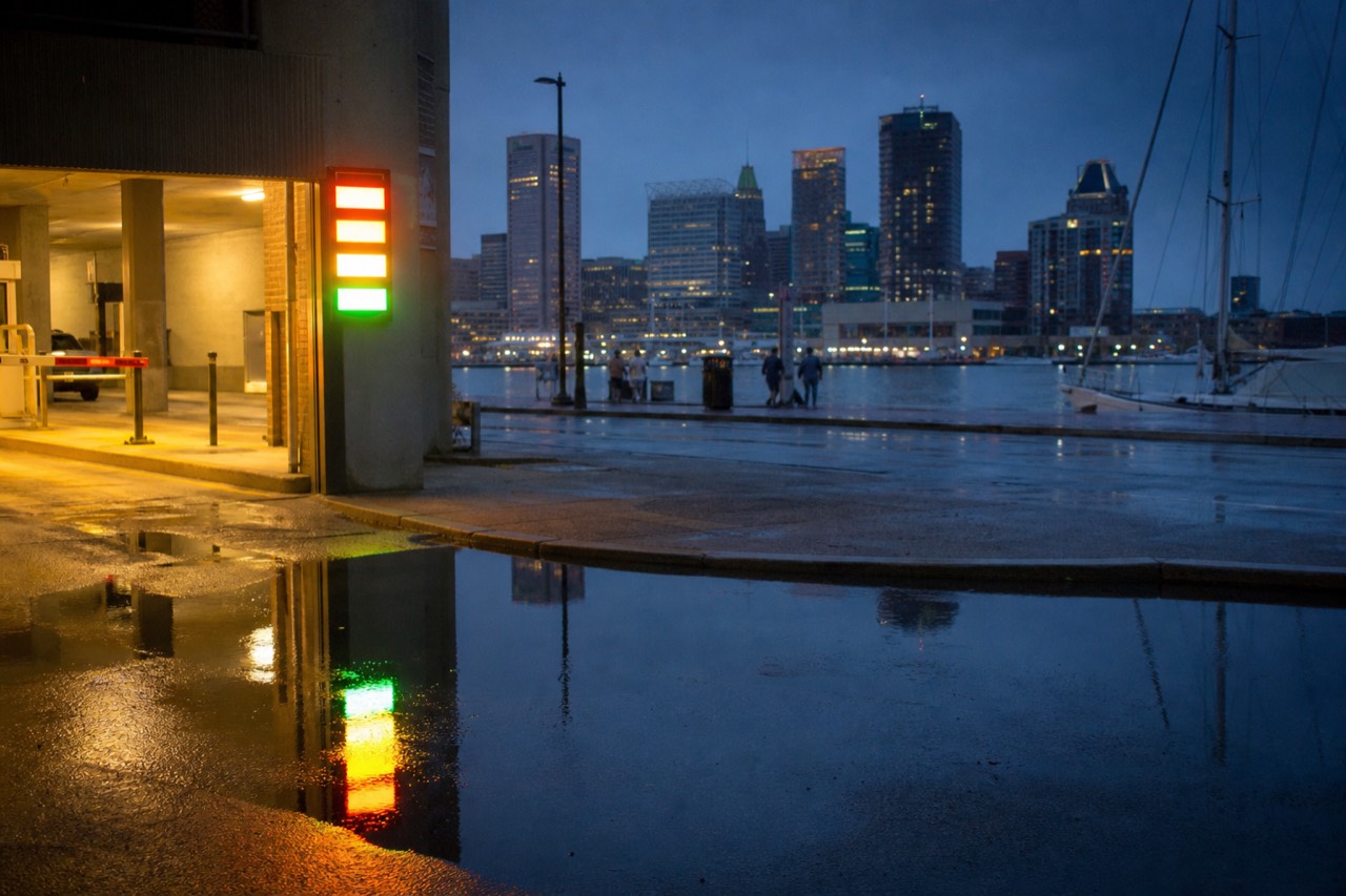 A rate board glowing at the mouth of a downtown parking structure at blue hour, the Inner Harbor water and skyline reflected in a puddle on the wet pavement