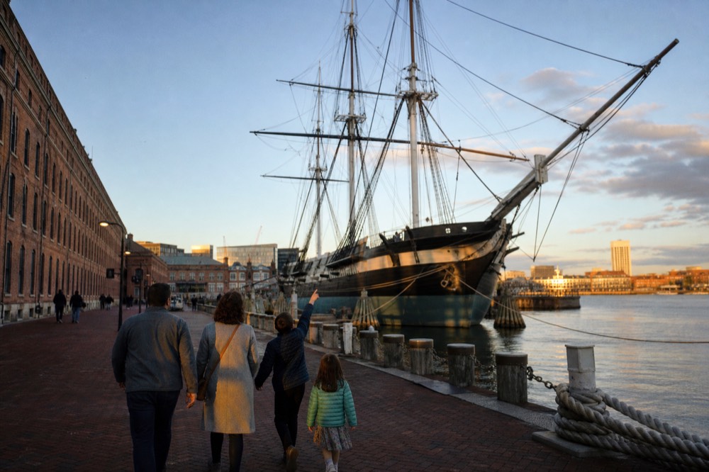 A family walking the brick waterfront promenade past a moored historic warship, kids pointing up at the tall masts under a blue sky