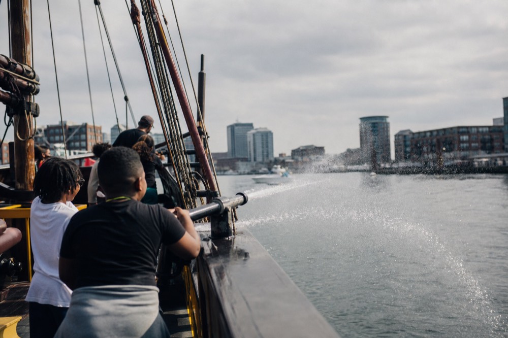 Kids laughing on the deck of a pirate-themed cruise boat spraying a water cannon toward the harbor, foam and spray catching the sun off Fells Point