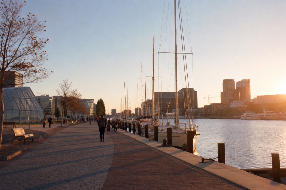 The 2.5-mile waterfront promenade curving along the harbor with benches and public art, people strolling past glass pavilions in the distance on a clear afternoon