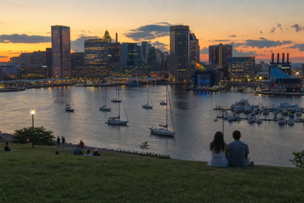 Federal Hill Park's grassy overlook with the full harbor and skyline spread below, a couple sitting on the slope at golden hour, warm light across the water