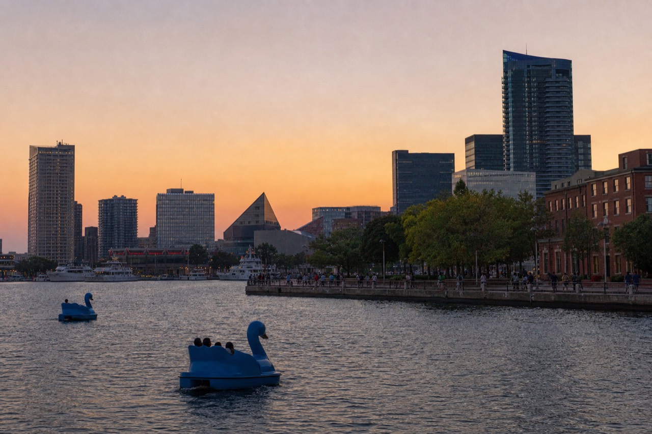 The Inner Harbor skyline and brick promenade seen from the water at midday, paddleboats on green water and families walking the waterfront under a bright blue sky