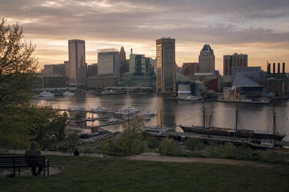Federal Hill Park at golden hour looking down over the Inner Harbor, the full skyline and moored ships spread out below a warm evening sky