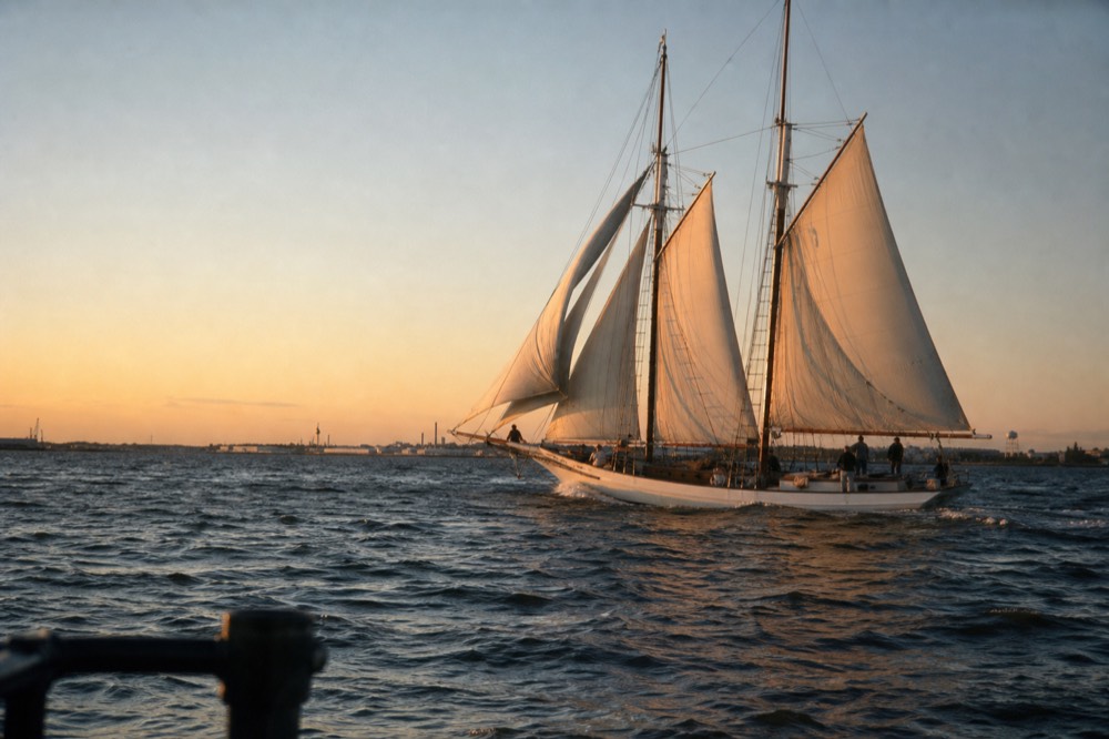 A white schooner heeling gently under full sail off Fort McHenry at sunset, the crew trimming lines, warm light low across the open Patapsco