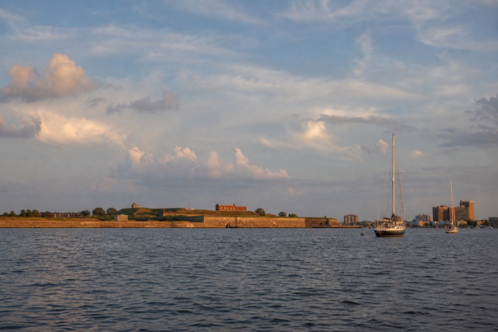 Fort McHenry's low star-shaped ramparts and green lawns seen from the water several miles down the Patapsco, a wide calm river under a big summer sky