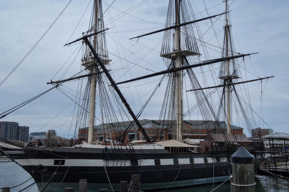 The masts and rigging of the USS Constellation historic sloop docked at the Inner Harbor, brick pavilions and a clear sky behind the tall wooden ship
