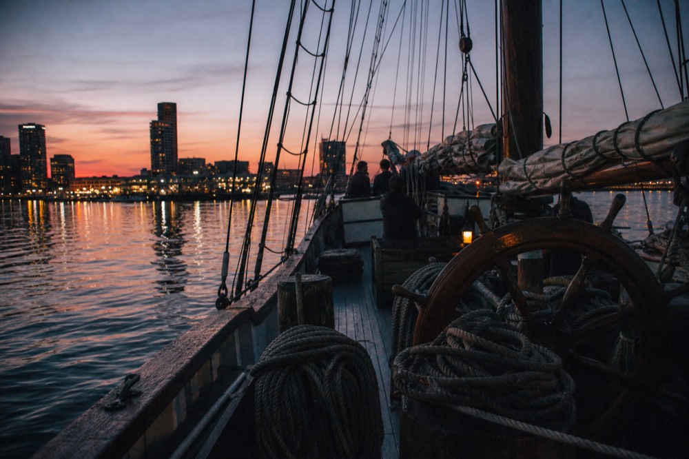 The open deck of a sailing schooner at blue hour, coiled ropes and a wooden wheel in the foreground, city lights reflecting on dark harbor water