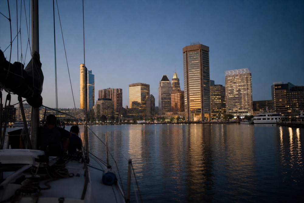 The Baltimore skyline seen from the water after sunset, office towers lit gold against a deep blue sky, the harbor calm and glassy