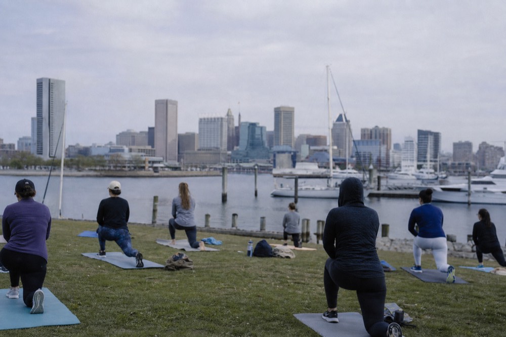 A morning outdoor fitness class on the waterfront grass beside the harbor, exercise mats laid out, the skyline and calm water in the background