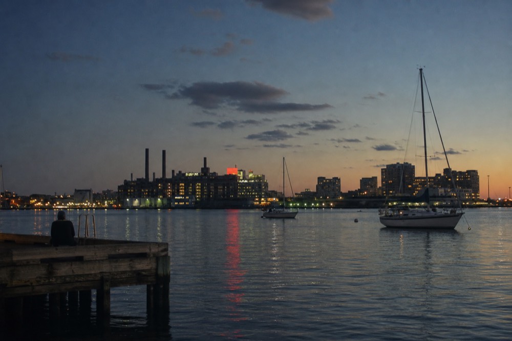 The Domino Sugars neon sign glowing red across the water at dusk, seen from Federal Hill, harbor lights reflecting on the dark surface
