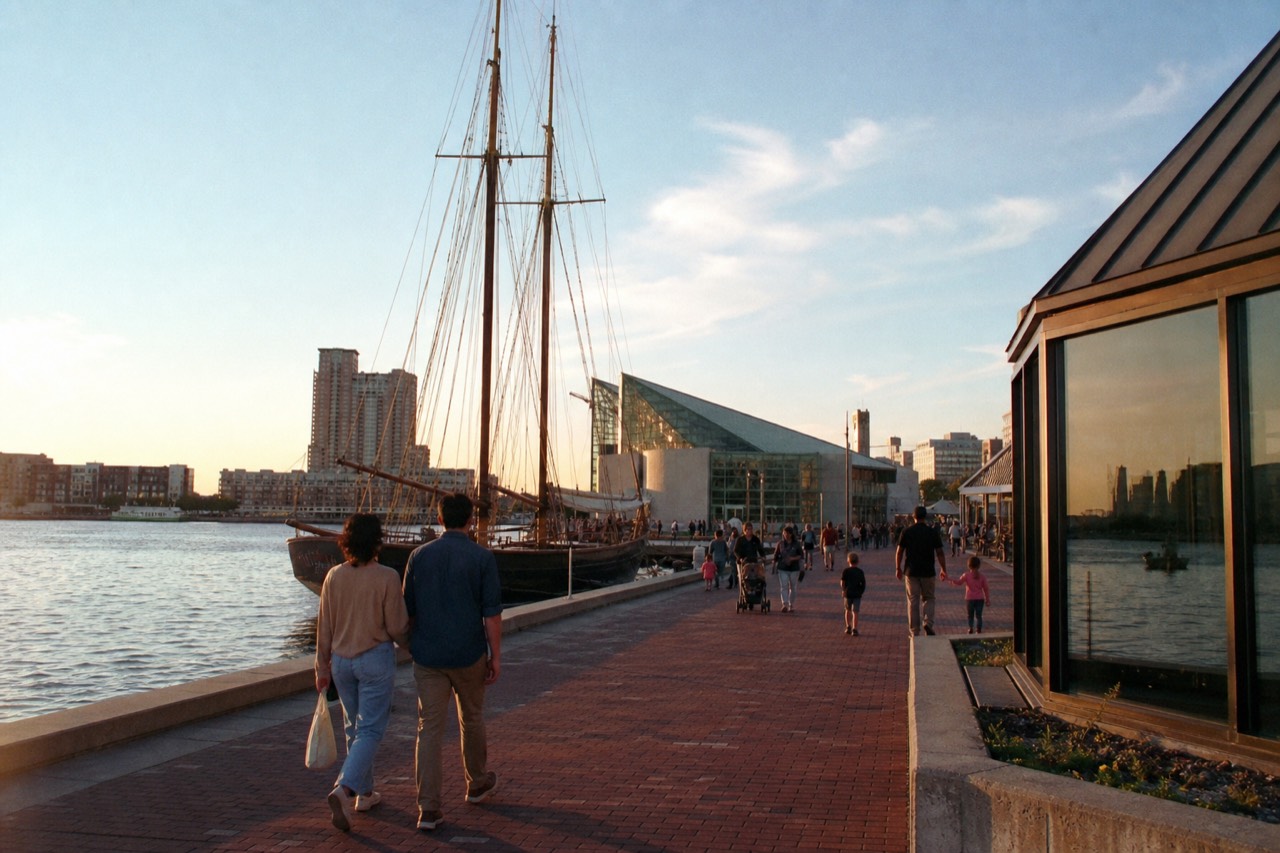 The Inner Harbor waterfront promenade on a bright morning, tall ship masts and glass aquarium pavilions along the brick walkway, families strolling by calm water