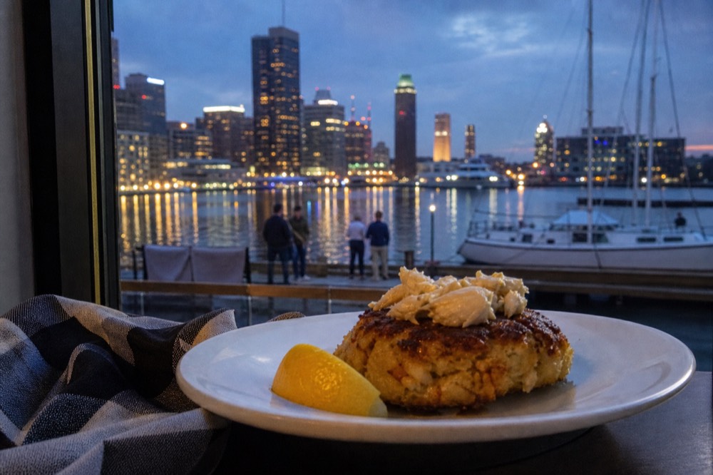 A single broiled crab cake mounded with lump crab meat on a simple white plate beside a lemon wedge, a checked cloth and a window of harbor light behind it