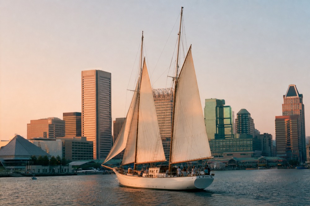 A white schooner under sail at golden hour on the Inner Harbor, the downtown skyline turning pink behind the tall masts and warm low light spread across calm water