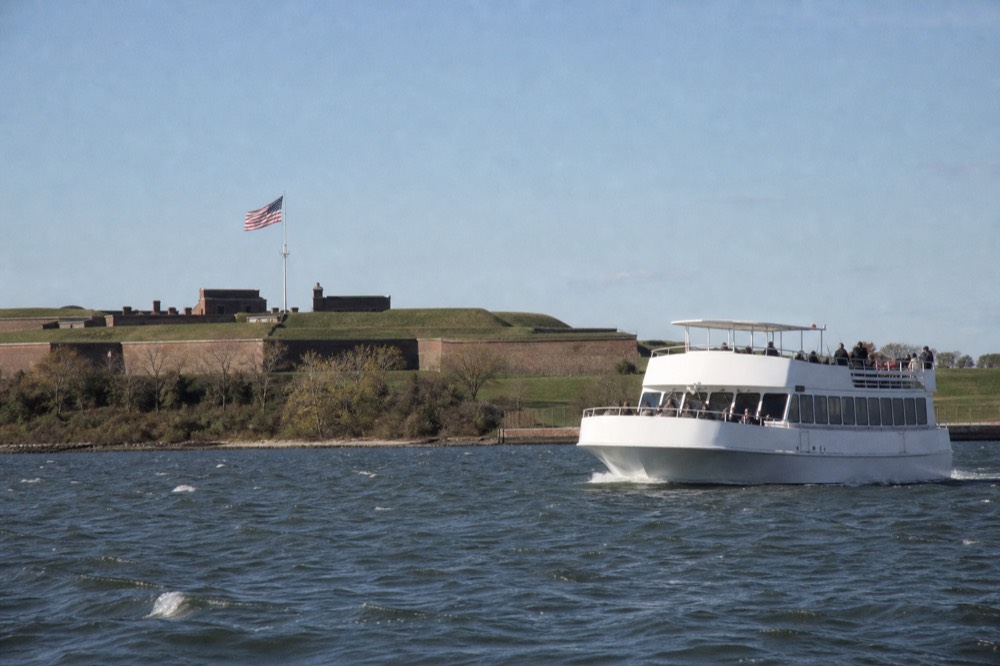A narrated white cruise boat gliding past Fort McHenry's star-shaped ramparts, the American flag flying over the fort, green lawns and calm water