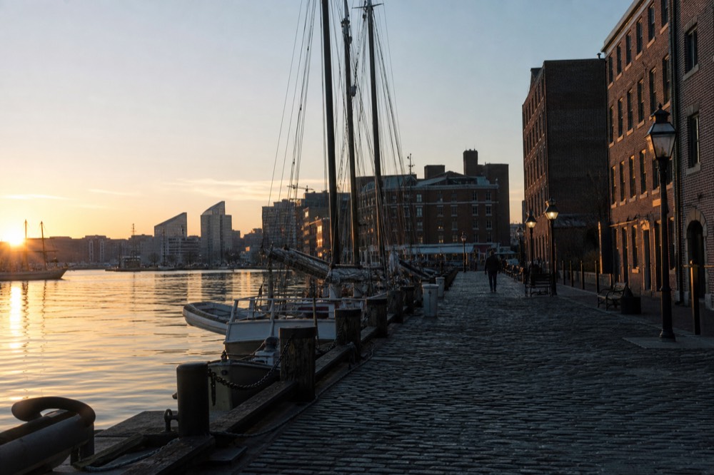 Fells Point's cobblestone waterfront at golden hour, tall-ship rigging and old brick warehouses, small boats tied along the pier