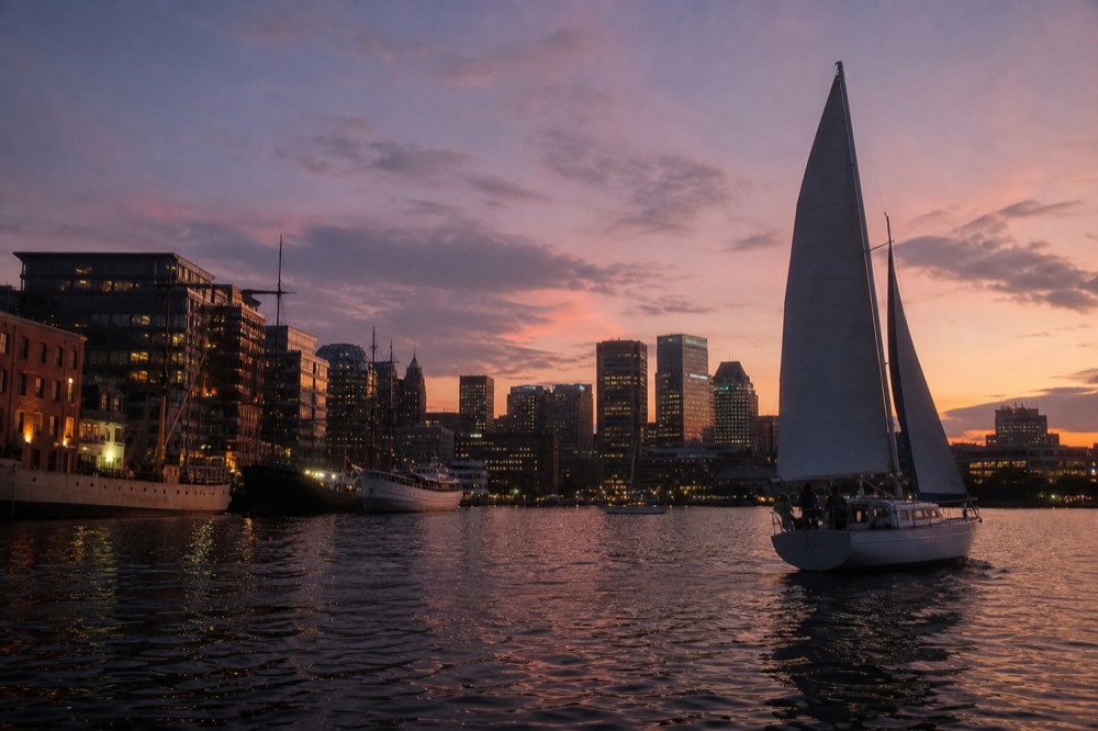 The Inner Harbor skyline lighting up at dusk from the water, office towers glowing warm, a sailboat silhouetted against a pink and violet sky