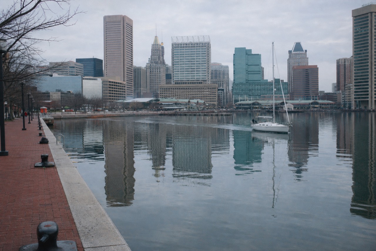 The Baltimore Inner Harbor at first light, still water reflecting the downtown towers, a single boat leaving a soft wake past the empty promenade
