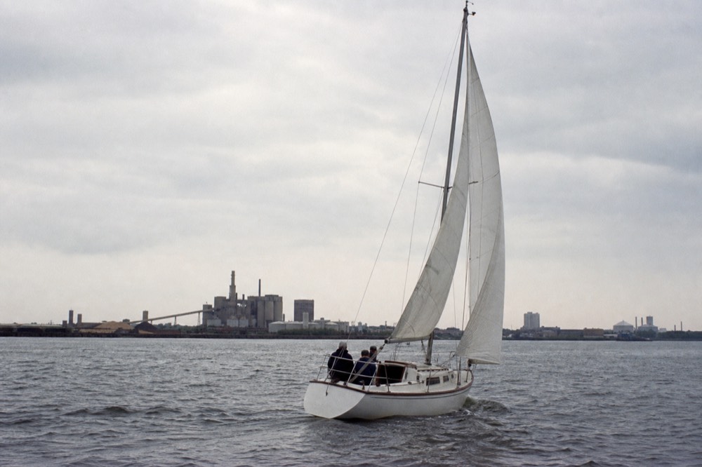 A white sailboat under working canvas leaning into a light breeze on the open Patapsco, the Domino Sugars refinery small on the far shore