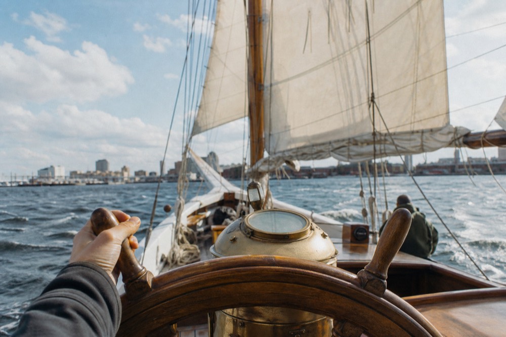 Close view of a schooner's wooden wheel and compass binnacle with the captain's hand resting on a spoke, sails full behind, harbor water sparkling