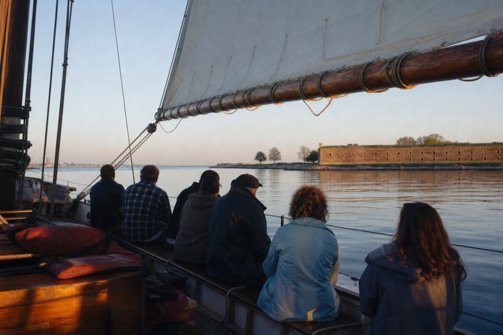 A small group of six seated along the rail of a traditional schooner under full sail, life jackets stowed on the bench, Fort McHenry low in the distance