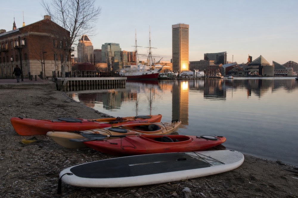 Kayaks and a paddleboard drawn up on a small urban launch beach beside the harbor promenade, calm morning water and the skyline mirrored in it
