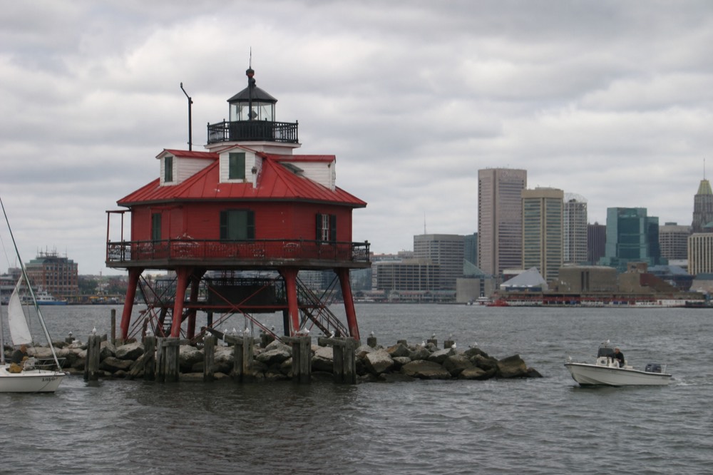 The Seven Foot Knoll Lighthouse, a squat red screw-pile light, seen from the water at Pier 5 with small boats passing and gulls on the pilings