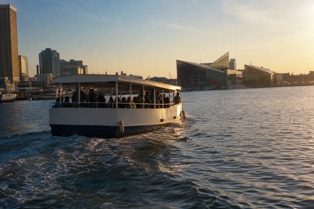 A covered narrated motor cruise boat mid-harbor with passengers standing along the rail, a wide wake fanning out behind it, glass waterfront pavilions in the hazy distance