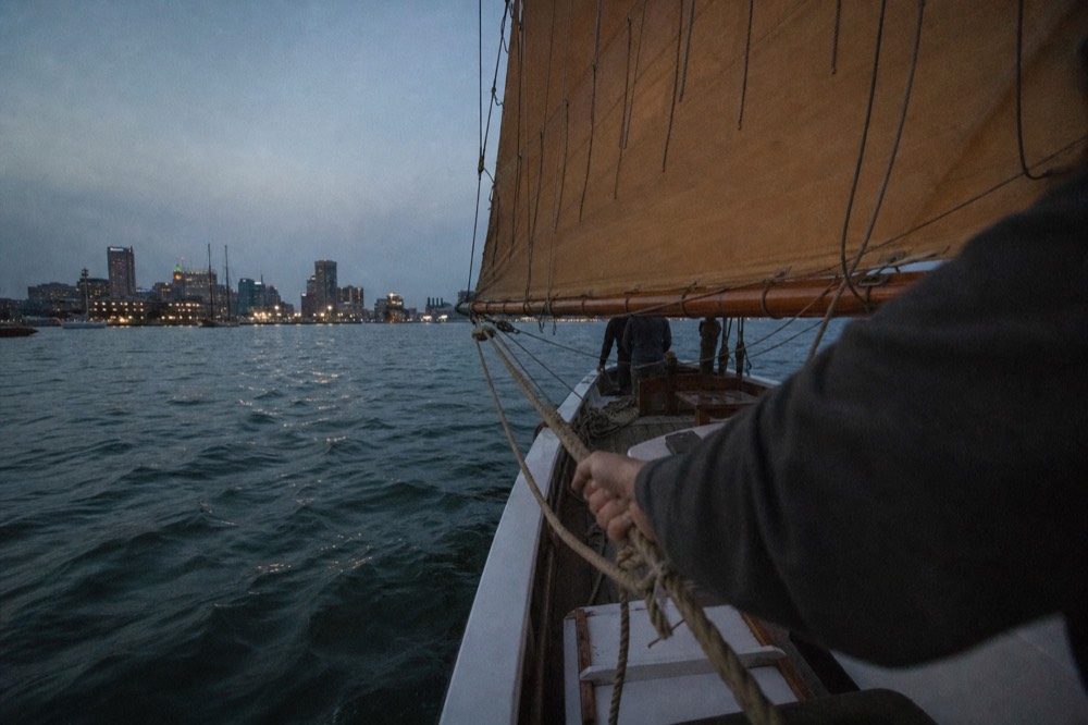 A traditional schooner's tan mainsail filled with wind, the boom swung out over green harbor water, a crew member's weathered hands easing a thick line
