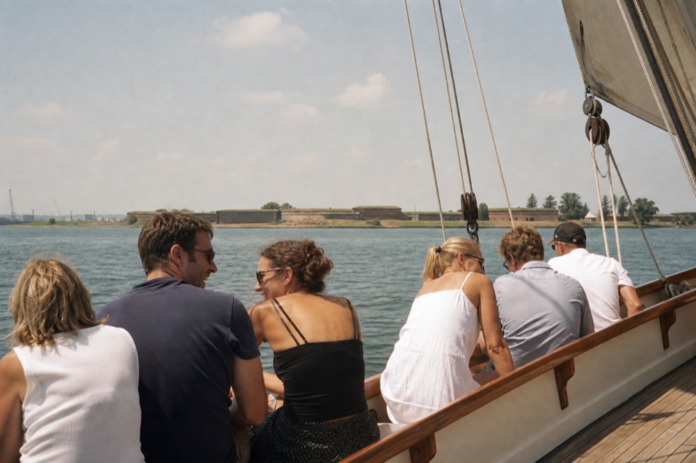 A small group of six seated along a schooner's rail laughing in the low sun, the distant brick ramparts of Fort McHenry across calm water behind them