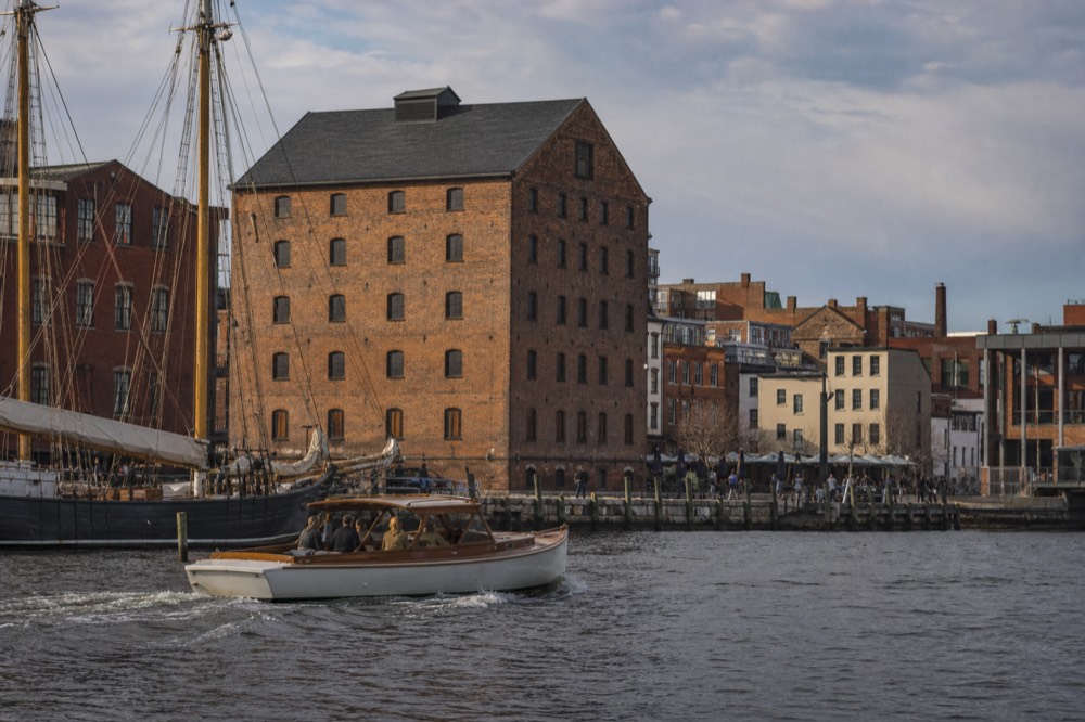 A small open harbor launch cutting across flat water past the cobblestone piers of Fells Point, brick warehouses and a tall ship's masts behind it in warm afternoon light