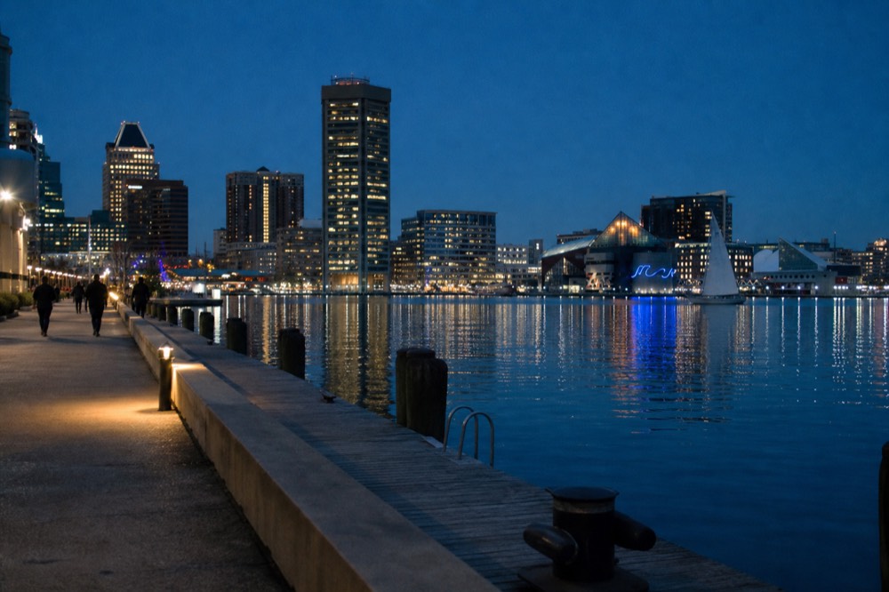Blue-hour city lights reflecting in long ribbons across calm harbor water, a single sailboat silhouette on the horizon under a deep indigo sky