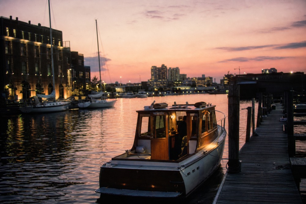 A small classic motor yacht idling at a wooden dock in the early evening, warm cabin lights glowing, brick warehouses and moored sailboats in the background