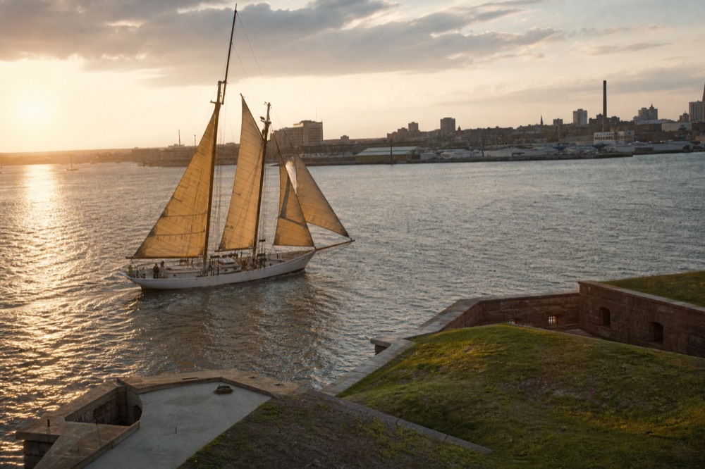 A traditional gaff-rigged schooner sailing close past the star-shaped ramparts of a waterfront fort, low evening sun gilding the sails and the grassy earthworks