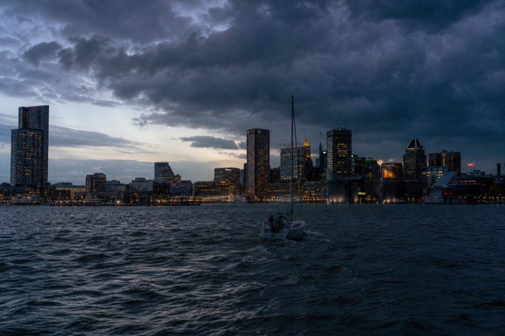 A summer storm building over the river in dark towering clouds, choppy grey water in the foreground and a small boat turning back toward the sheltered harbor
