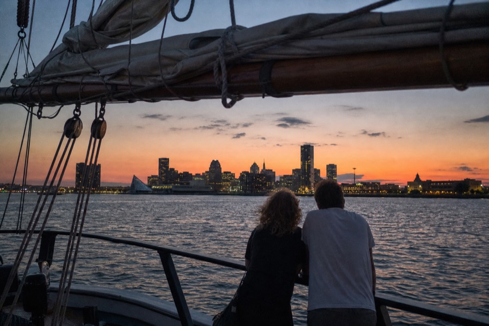Two people at the rail of a schooner watching the last orange light fade over the water, silhouetted rigging above them and the first city lights coming on across the harbor