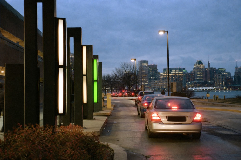 A row of illuminated garage rate signs on a downtown street near the waterfront in the evening, cars queuing at a ticket gate under a darkening sky