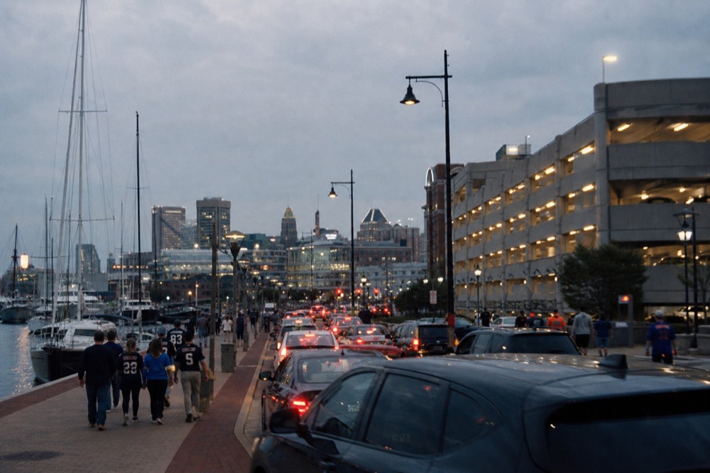 A busy downtown street near the harbor on a game evening, brake lights lined up toward a full garage and pedestrians in team colors crossing under streetlights