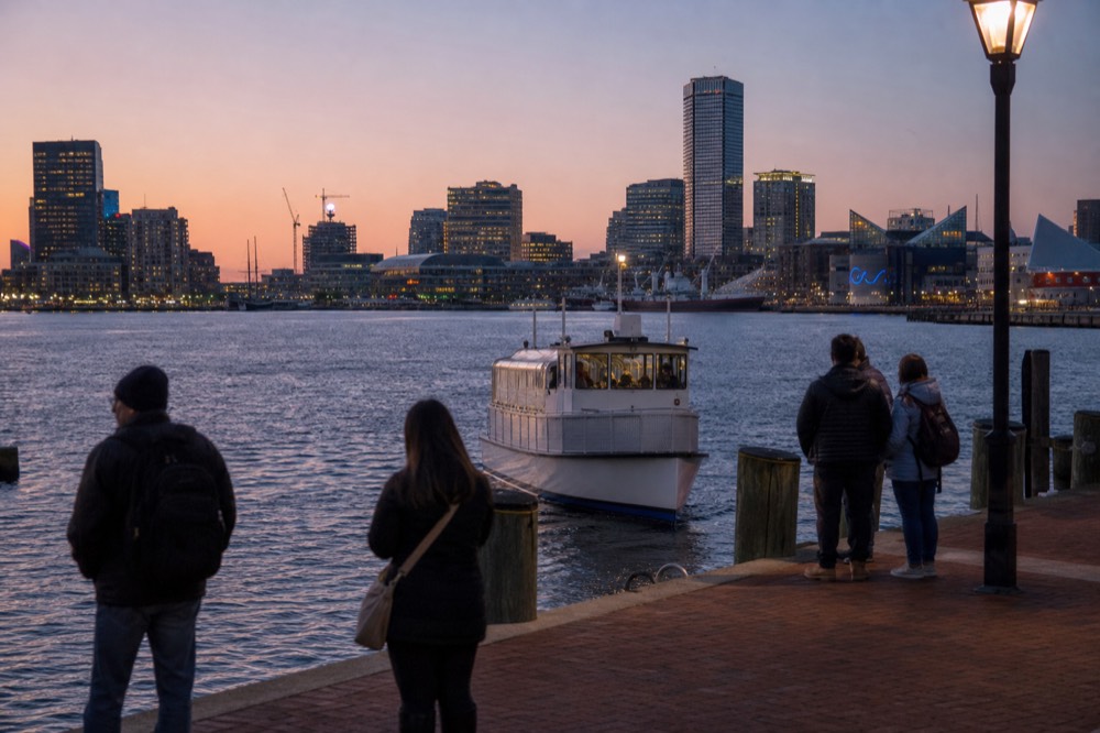 A small white passenger launch approaching a harbor dock with the skyline behind, a few riders waiting on the boardwalk in soft late-day light