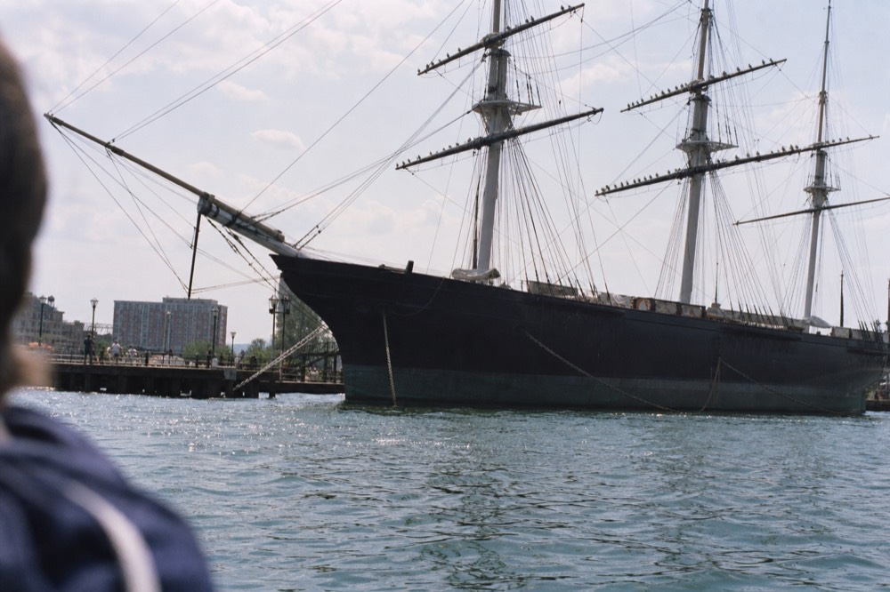 The black hull and tall rigging of a museum warship at a pier seen from a passing boat, gulls perched along the yardarms, soft morning light on the water