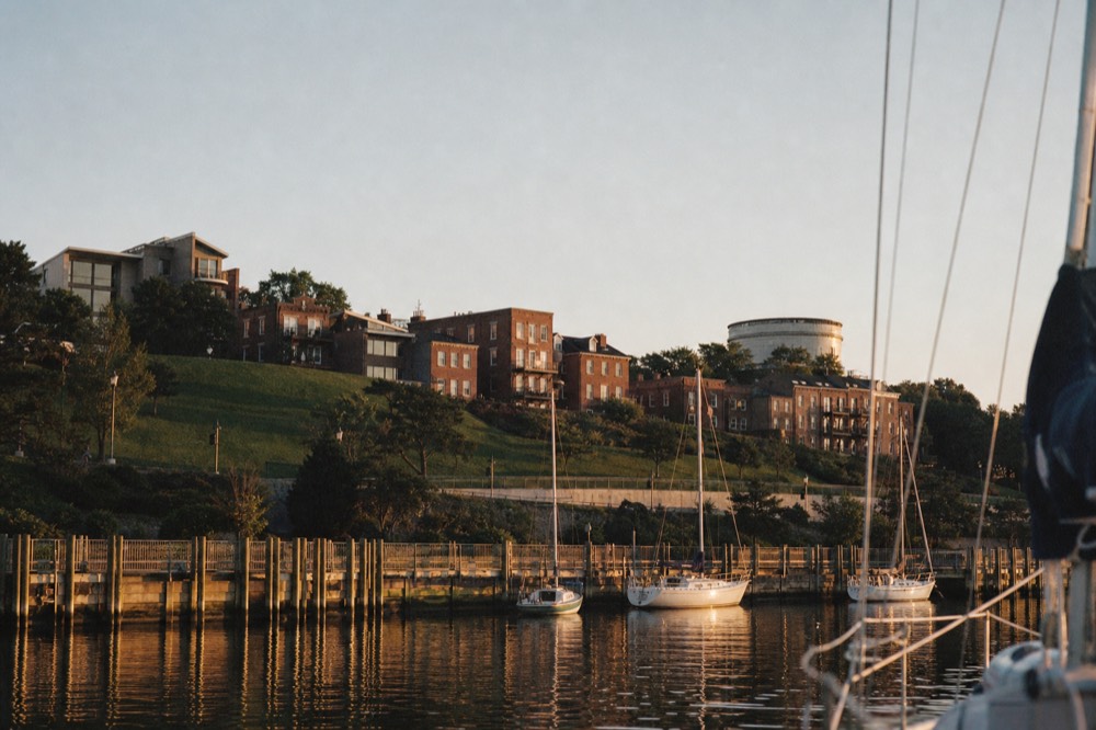 The green slope and brick rowhouses of Federal Hill rising over the south shore of the harbor, viewed low from a boat on calm water in the late afternoon