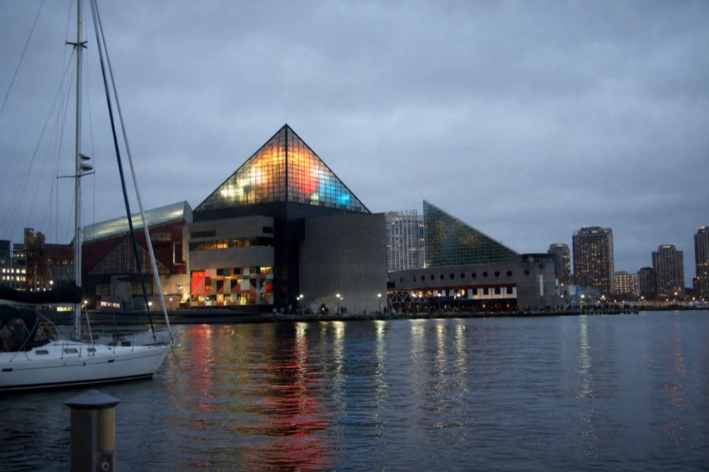 The illuminated glass pavilions of the National Aquarium seen from the water at nightfall, colored light spilling onto the promenade and the harbor surface