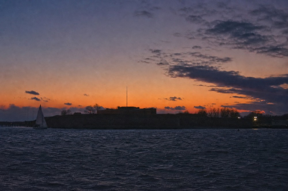 Fort McHenry's low ramparts and flagpole silhouetted against a deep orange and purple afterglow, the wide Patapsco going dark in the foreground