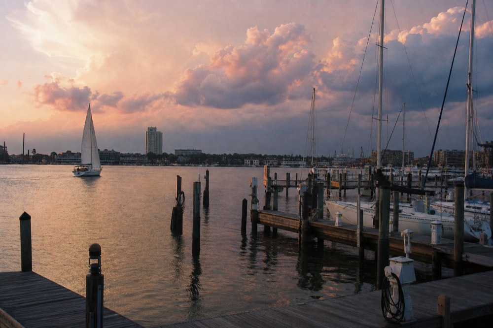 A summer thunderhead piling up pink and grey over the harbor at sunset, a distant sailboat turning back toward the docks under a bruised sky