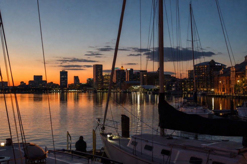 Blue-hour reflections of the Baltimore skyline stretching across glassy harbor water, boat masts in the foreground and the first stars overhead